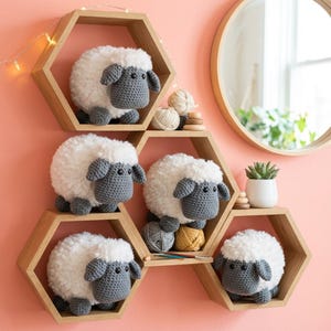 May include: Five handmade crochet sheep with white fluffy bodies and gray faces and legs are displayed on wooden hexagon shelves. The shelves are mounted on a pink wall, with a round mirror and a small potted plant visible in the background.