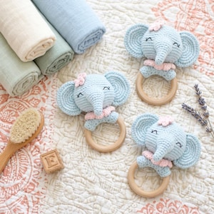 May include: Three light blue crochet elephant baby rattles with wooden rings and pink accents. Rolled pastel-colored baby blankets and a wooden baby brush are also visible. The rattles have a flower on their heads.