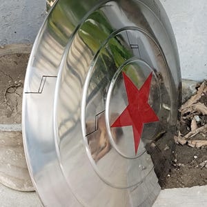 May include: A polished, metallic shield with a red star emblem at its center. The shield has a circular design with concentric rings and black accent lines. The surface reflects the surrounding environment.