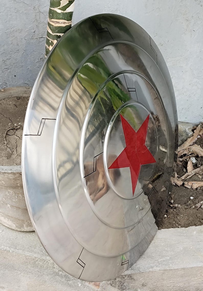 May include: A polished, metallic shield with a red star emblem at its center. The shield has a circular design with concentric rings and black accent lines. The surface reflects the surrounding environment.