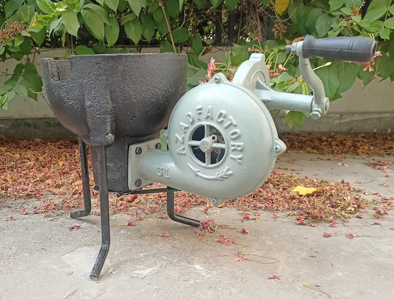 May include: A black cast iron forge with a hand-crank bellows. The bellows is painted light blue and has the text "AZAD FACTORY" on it. The forge is on a set of three black metal legs.