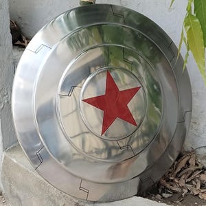 May include: A silver-colored, circular shield with a prominent red star in the center. The shield has concentric rings and black geometric designs around the edges, giving it a futuristic look. The shield is likely made of metal.