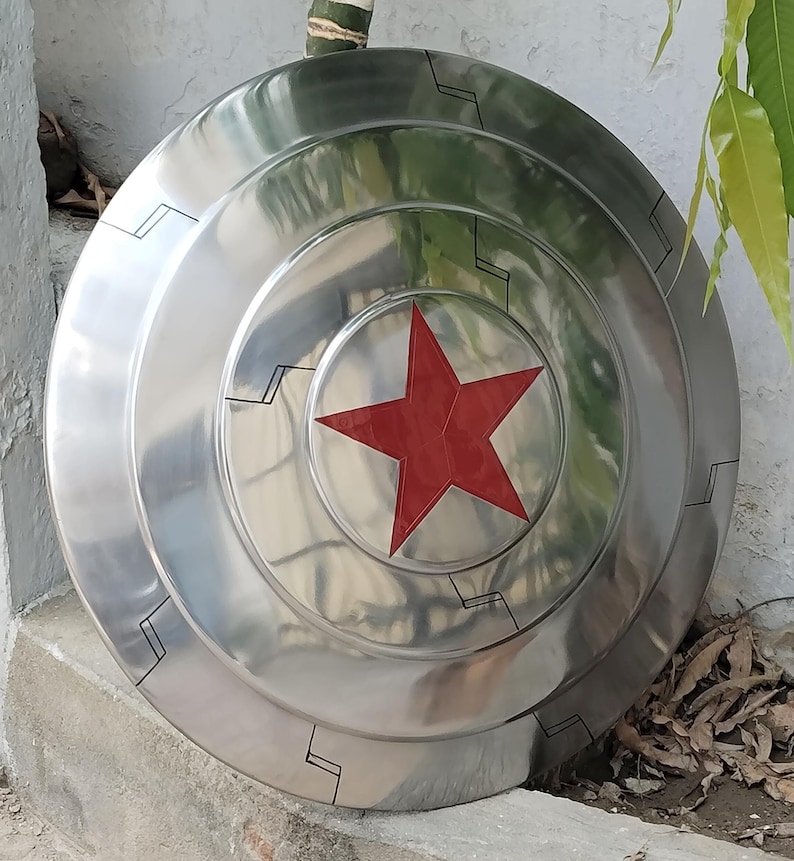 May include: A silver-colored, circular shield with a prominent red star in the center. The shield has concentric rings and black geometric designs around the edges, giving it a futuristic look. The shield is likely made of metal.