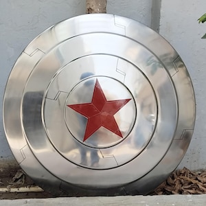 May include: A silver-colored, circular shield with a prominent red star in the center. The shield has concentric rings and black geometric designs. The shield is a prop or collectible item.