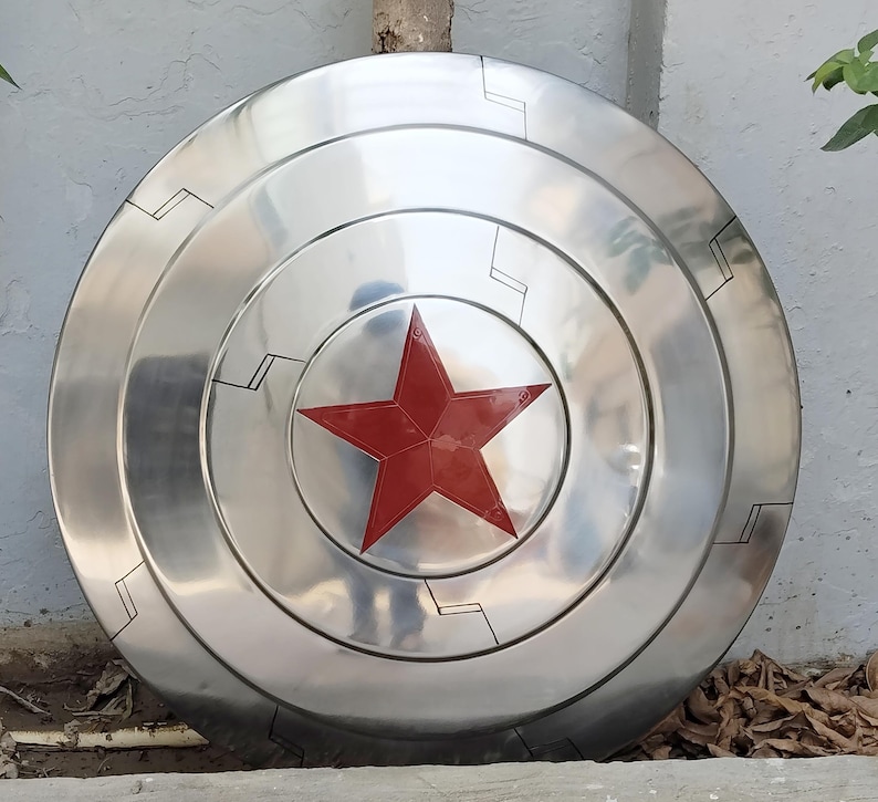 May include: A silver-colored, circular shield with a prominent red star in the center. The shield has concentric rings and black geometric designs. The shield is a prop or collectible item.