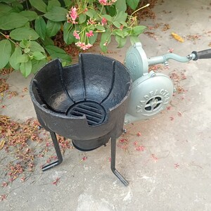 May include: A black metal forge with a round bowl and three legs. A gray hand-crank bellows with a black handle is attached to the side of the forge.
