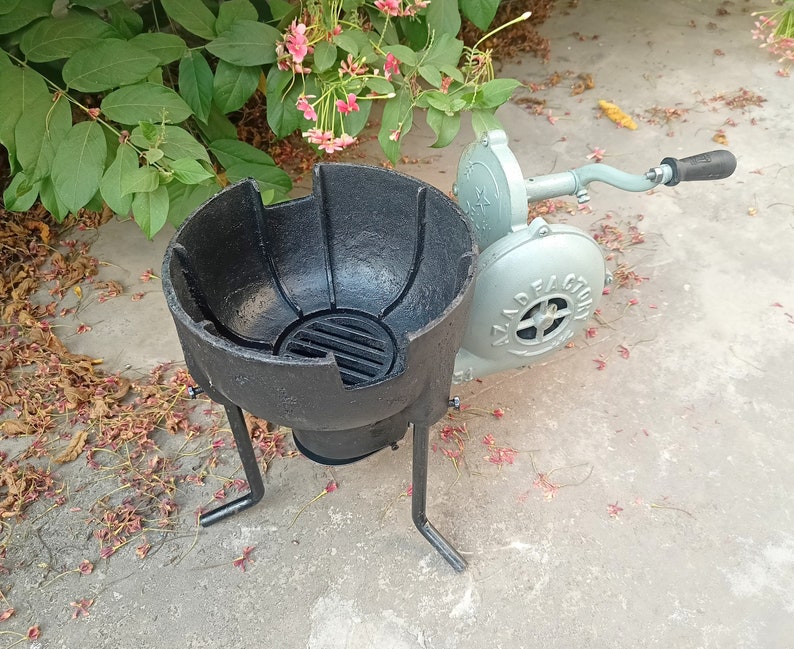 May include: A black metal forge with a round bowl and three legs. A gray hand-crank bellows with a black handle is attached to the side of the forge.