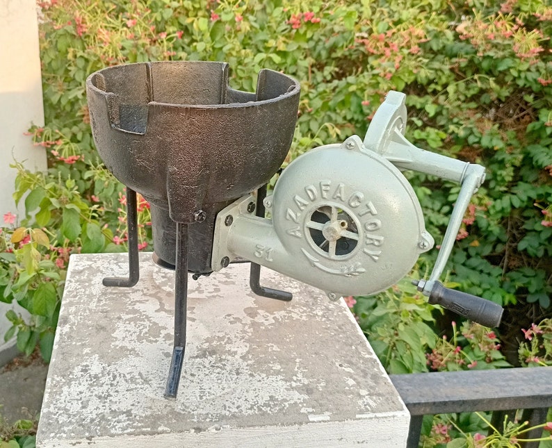 May include: A black cast iron stove with three legs and a hand-crank bellows attached. The bellows are painted light gray with the text "AZAD FACTORY" printed on the side.