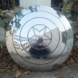 May include: A shiny, silver shield with a five-pointed star in the center. The shield has concentric circular ridges and a reflective surface. The shield is a prop or collectible item.