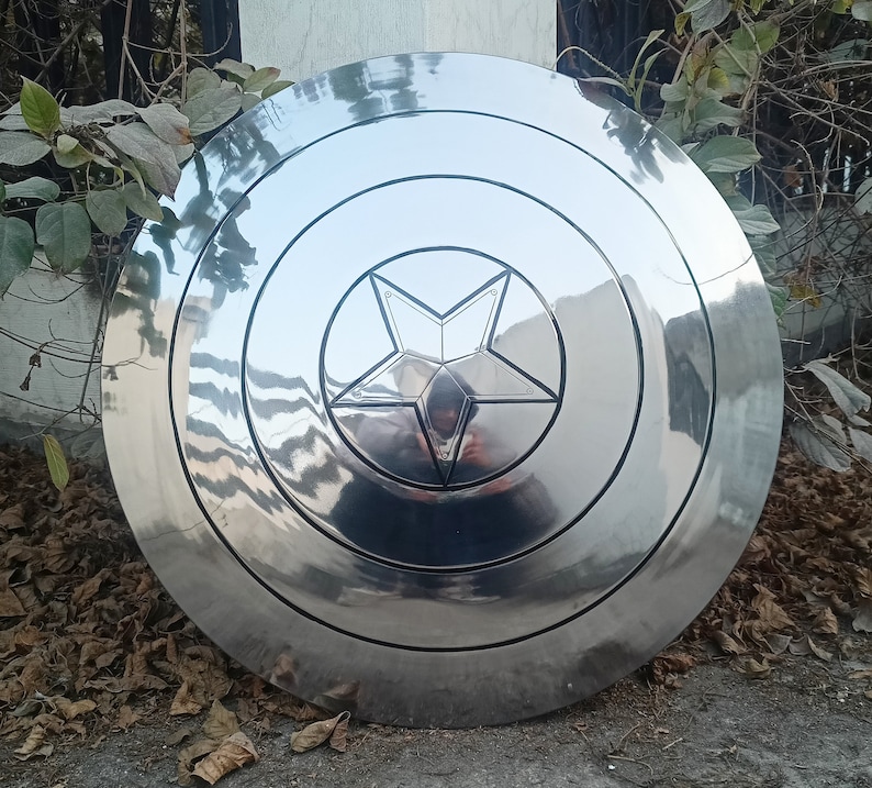 May include: A shiny, silver shield with a five-pointed star in the center. The shield has concentric circular ridges and a reflective surface. The shield is a prop or collectible item.