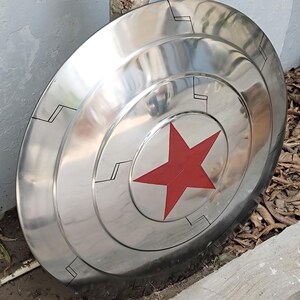 May include: A polished, metallic shield with a red star at the center. The shield has concentric circular bands and black geometric designs. The overall design is reminiscent of a superhero's iconic shield.