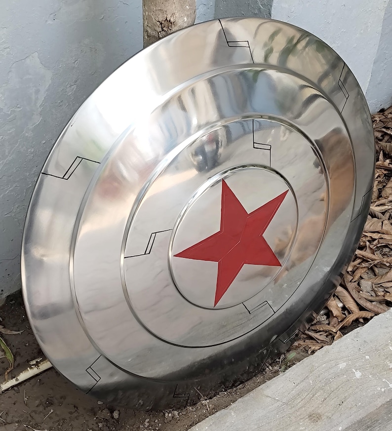 May include: A polished, metallic shield with a red star at the center. The shield has concentric circular bands and black geometric designs. The overall design is reminiscent of a superhero's iconic shield.