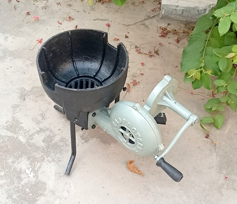 May include: A black cast iron forge with a blower attached. The blower is silver and has a handle. The forge is on a concrete surface.