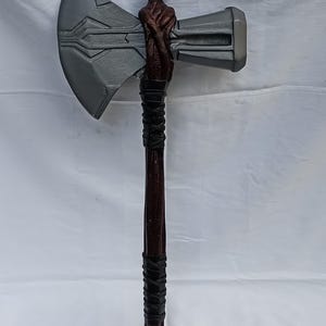 May include: A large, gray and brown battle axe with a detailed design on the blade. The axe head is gray, and the handle is dark brown with black leather wrapping. The axe is a prop or decorative item.