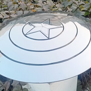 May include: A silver-colored, circular shield with a central five-pointed star design. The shield has concentric circular grooves and a reflective surface, reminiscent of a superhero's iconic accessory. The shield is a prop or collectible.