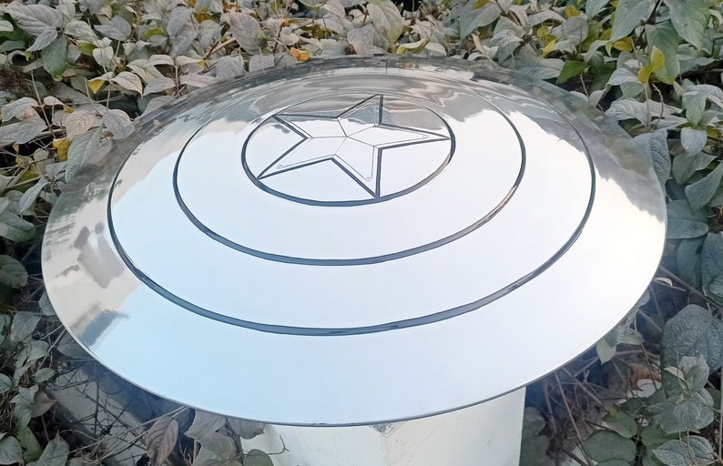 May include: A silver-colored, circular shield with a central five-pointed star design. The shield has concentric circular grooves and a reflective surface, reminiscent of a superhero's iconic accessory. The shield is a prop or collectible.