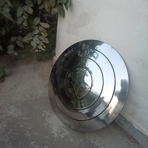 May include: A polished, metallic shield with a central star design. The shield is circular with concentric rings and a five-pointed star in the center. The surface reflects light, giving it a shiny appearance. The shield is leaning against a white wall.