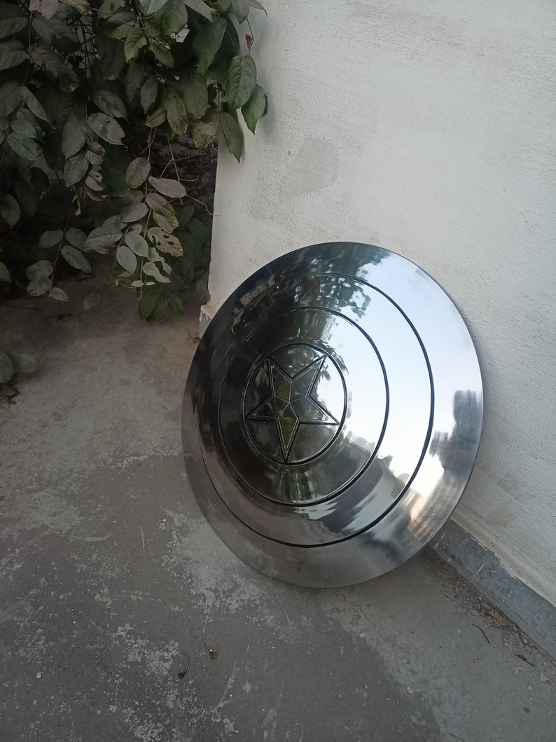 May include: A polished, metallic shield with a central star design. The shield is circular with concentric rings and a five-pointed star in the center. The surface reflects light, giving it a shiny appearance. The shield is leaning against a white wall.