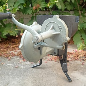 May include: A gray metal hand-crank grain mill with a black metal hopper and a black metal stand. The mill has a handle and a crank that turns a set of gears.
