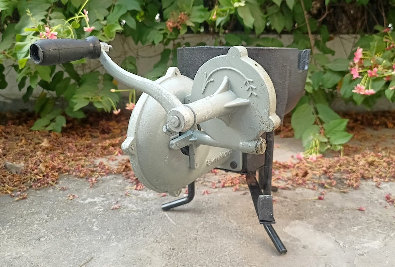 May include: A gray metal hand-crank grain mill with a black metal hopper and a black metal stand. The mill has a handle and a crank that turns a set of gears.