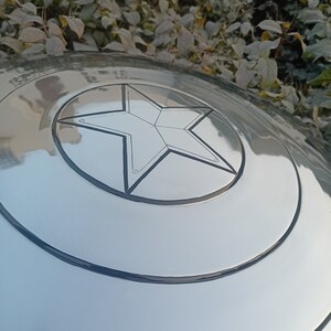 May include: A silver-colored shield with a prominent five-pointed star emblem at its center. The shield has concentric circular bands and a reflective surface, suggesting a metallic construction. The star is outlined in black.
