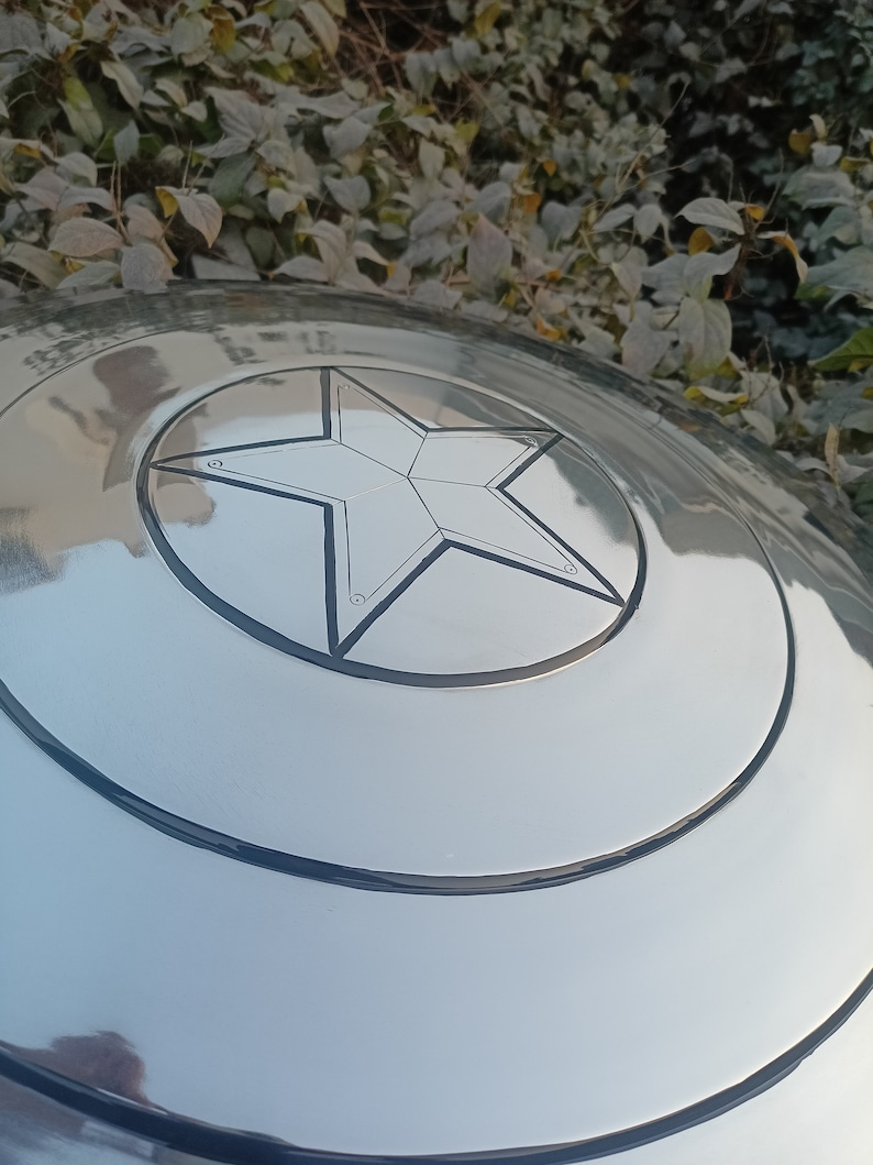 May include: A silver-colored shield with a prominent five-pointed star emblem at its center. The shield has concentric circular bands and a reflective surface, suggesting a metallic construction. The star is outlined in black.