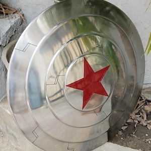 May include: A silver-colored shield with a red star in the center. The shield has concentric circular ridges and black geometric markings. The shield is a prop or collectible item.