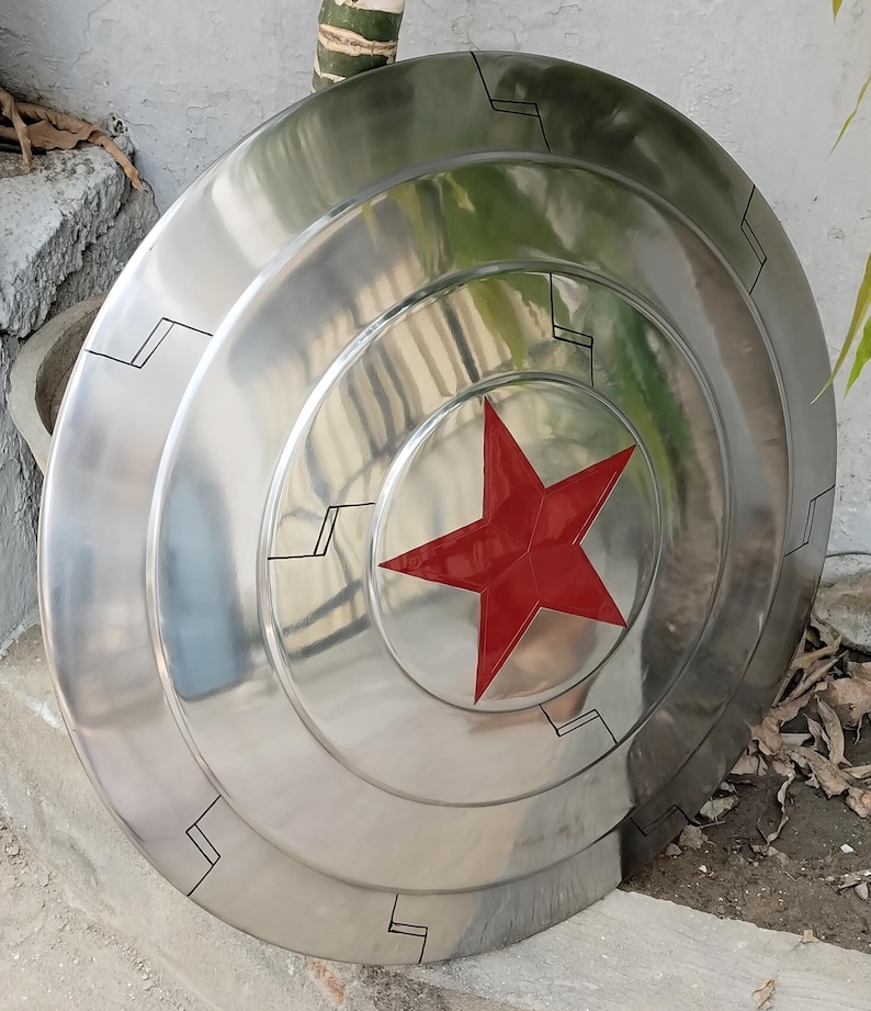 May include: A silver-colored shield with a red star in the center. The shield has concentric circular ridges and black geometric markings. The shield is a prop or collectible item.