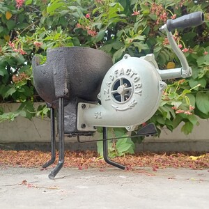May include: A black metal forge with a gray hand-crank blower. The blower has the text "AZAD FACTORY" printed on it. The forge is on a concrete surface with fallen leaves.