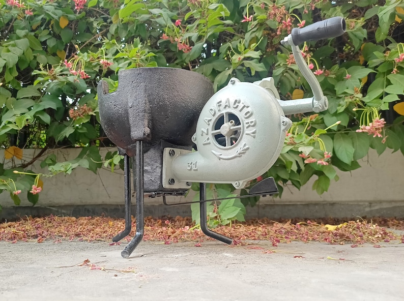 May include: A black metal forge with a gray hand-crank blower. The blower has the text "AZAD FACTORY" printed on it. The forge is on a concrete surface with fallen leaves.