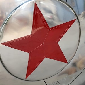 May include: A silver-colored shield with a large, red, five-pointed star in the center. The star is outlined and has a glossy finish. The shield has a circular shape with a reflective surface.