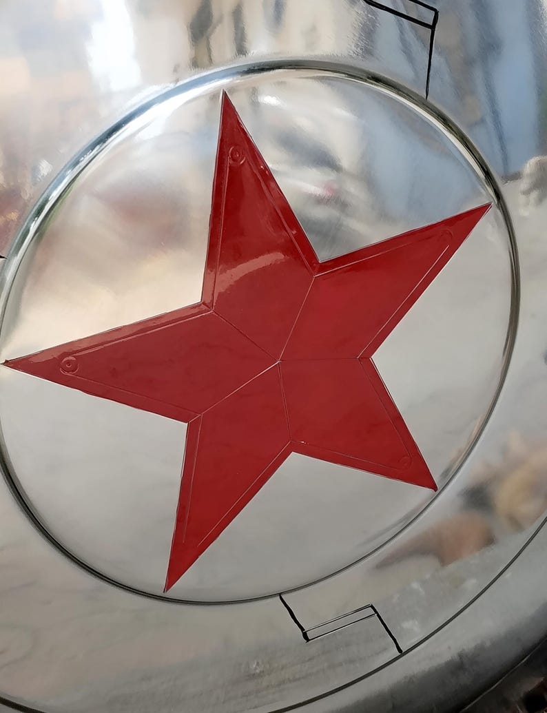 May include: A silver-colored shield with a large, red, five-pointed star in the center. The star is outlined and has a glossy finish. The shield has a circular shape with a reflective surface.