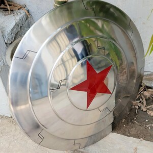 May include: A polished, metallic shield with a red star at the center. The shield has concentric circular ridges and black geometric accents. The overall design is reminiscent of a superhero's iconic shield.
