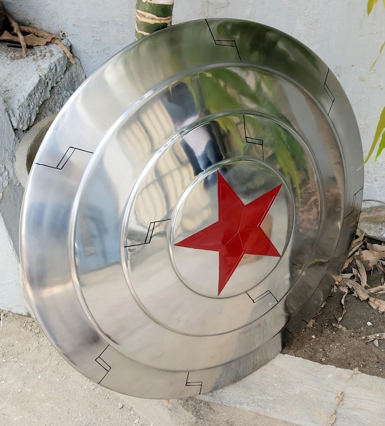 May include: A polished, metallic shield with a red star at the center. The shield has concentric circular ridges and black geometric accents. The overall design is reminiscent of a superhero's iconic shield.