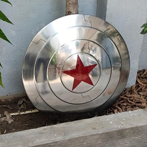 May include: A polished, metallic shield with a central red star emblem. The shield has concentric circular ridges and etched linear details. The overall design is a classic, iconic superhero accessory, reflecting a theme of protection and heroism.