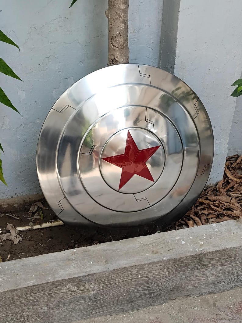 May include: A polished, metallic shield with a central red star emblem. The shield has concentric circular ridges and etched linear details. The overall design is a classic, iconic superhero accessory, reflecting a theme of protection and heroism.