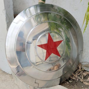 May include: A silver-colored shield with a prominent red star in the center. The shield has concentric circular ridges and black geometric designs. The surface reflects light, giving it a metallic appearance. A prop or collectible.