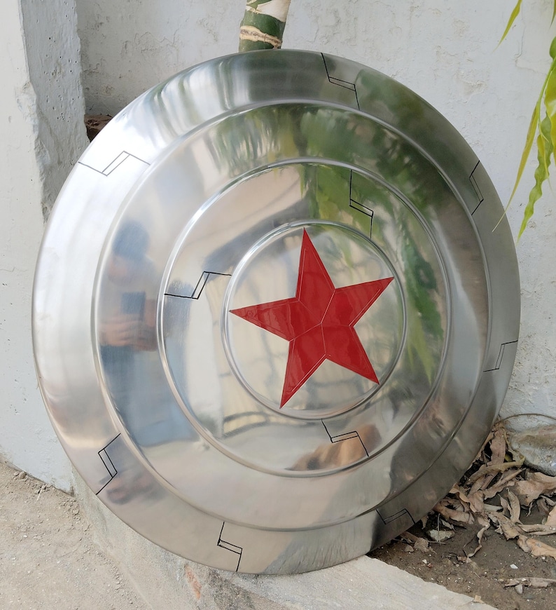 May include: A silver-colored shield with a prominent red star in the center. The shield has concentric circular ridges and black geometric designs. The surface reflects light, giving it a metallic appearance. A prop or collectible.