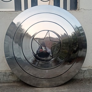 May include: A polished, metallic shield with a central five-pointed star emblem. The shield is circular, with concentric rings and a reflective surface. The star is recessed and outlined, adding depth to the design. A classic superhero accessory.