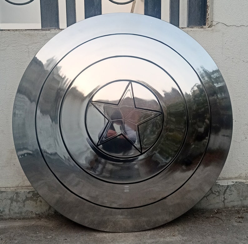 May include: A polished, metallic shield with a central five-pointed star emblem. The shield is circular, with concentric rings and a reflective surface. The star is recessed and outlined, adding depth to the design. A classic superhero accessory.
