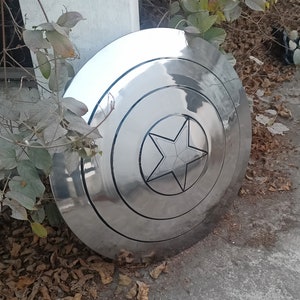 May include: A large, shiny, silver shield with a prominent five-pointed star in the center. The shield has concentric circular bands and a polished, reflective surface. The star is outlined in black, and the shield appears to be made of metal.