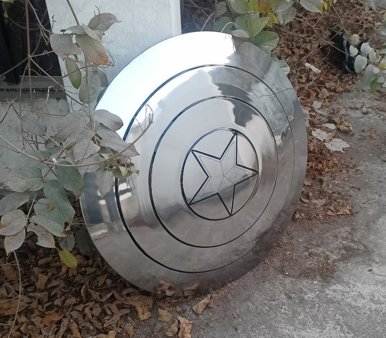May include: A large, shiny, silver shield with a prominent five-pointed star in the center. The shield has concentric circular bands and a polished, reflective surface. The star is outlined in black, and the shield appears to be made of metal.