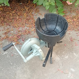 May include: A black cast iron charcoal brazier with a grill grate and three legs. A hand-crank bellows with a white metal body is attached to the brazier.