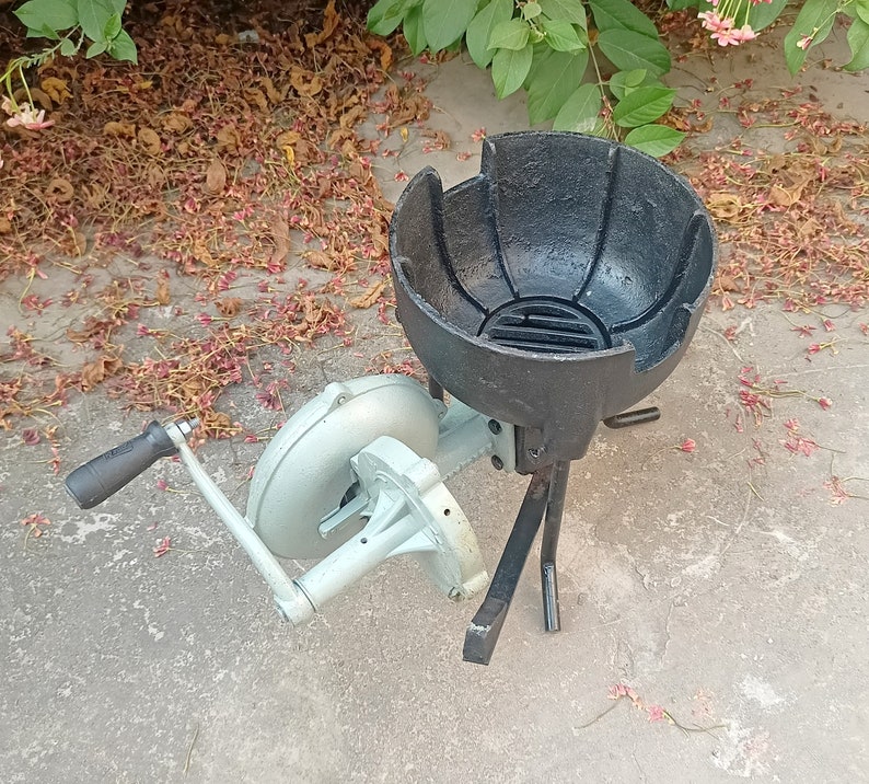 May include: A black cast iron charcoal brazier with a grill grate and three legs. A hand-crank bellows with a white metal body is attached to the brazier.