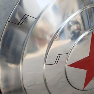 May include: A metallic silver shield with a red star emblem. The shield has concentric circular ridges and black accent lines. The surface reflects light, creating a shiny appearance. A classic superhero accessory.