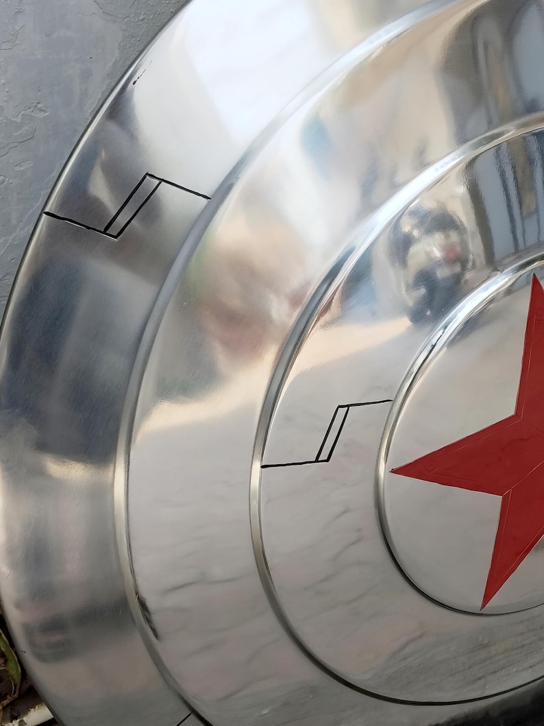 May include: A metallic silver shield with a red star emblem. The shield has concentric circular ridges and black accent lines. The surface reflects light, creating a shiny appearance. A classic superhero accessory.