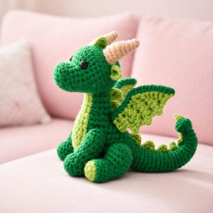 May include: A handmade crochet dragon in shades of green, with lighter green wings and accents. The dragon has small black eyes and tan horns. It is sitting on a pink surface.