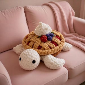 Op de afbeelding: Een gehaakte schildpad knuffel met een taartvormige schild. De schild is bruin met een rasterpatroon, bekroond met witte slagroom, een rode aardbei en blauwe bessen. De kop en ledematen van de schildpad zijn wit.