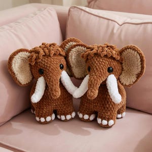 May include: Two brown crochet mammoth plush toys with cream-colored tusks, ears, and feet. The plushies have black eyes and are positioned close together. The background is a pink sofa.
