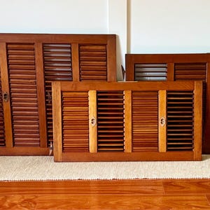 May include: A collection of vintage wooden shutters in various sizes, leaning against a white wall. The shutters are a warm brown color with horizontal slats and metal hardware. They rest on a beige rug and a wooden floor.